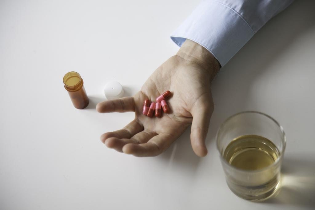 Ein Mann hält in seiner Hand fünf rosarote Schlaftabletten. Daneben stehen ein offenes Tablettenröhrchen und ein Glas Alkohol.
