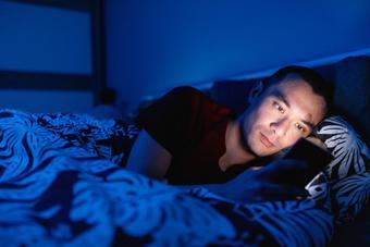 Mixed race man using smart phone in bed next to his sleeping partner