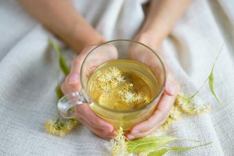 A cup of herbal tea (lime blossom/ linden tea) photographed on a cozy beige blanket with herbal flowers in the background.